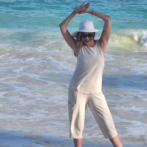 Person in white outfit and hat stretching on a beach with blue water.