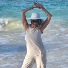 Load image into Gallery viewer, Woman in a white hat and sleeveless top standing on a beach with ocean waves in the background