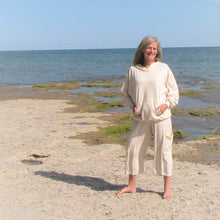 Load image into Gallery viewer, A person standing on a beach with capris on, facing the ocean and rocks in the foreground.