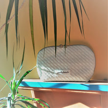 Load image into Gallery viewer, white makeup bag sitting on a shelf. Plant leaves are around it 